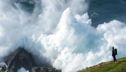 Hiker standing on grassy cliff watching massive ocean waves crash against dark rock formations.
