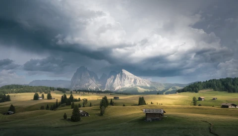 Alpine mountain valley with dramatic storm clouds, scattered farmhouses, and forested slopes beneath towering peaks.