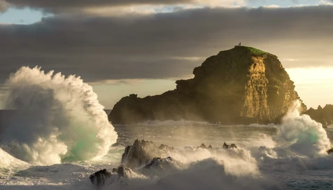 Large ocean waves crash against a rocky sea stack during golden hour with dramatic storm clouds overhead.