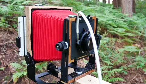 Large format camera with red bellows mounted on wooden frame in forest setting.