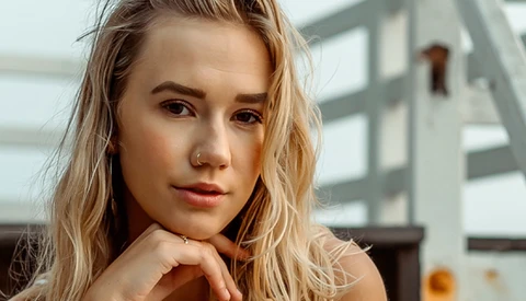Young woman with blonde wavy hair resting her chin on her hand, gazing directly at the camera with a contemplative expression.