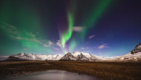 Northern lights dancing in green and purple bands above snow-capped mountains and a frozen lake.