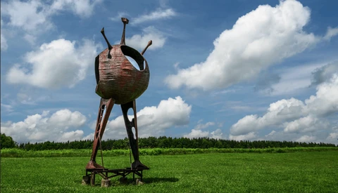 Rusted metal sculptural vessel with three protruding legs standing alone in a vast green field under cloudy blue sky.