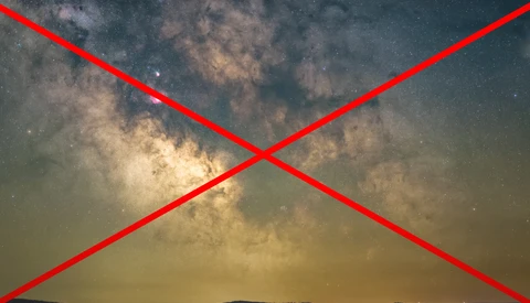 Milky Way galaxy stretching across night sky above dark mountain silhouette with red X overlay.