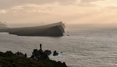 Silhouetted figure standing on rocky coastline overlooking a tilted shipwreck in misty waters.