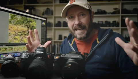Photographer gesturing expressively while discussing camera equipment displayed on a table.