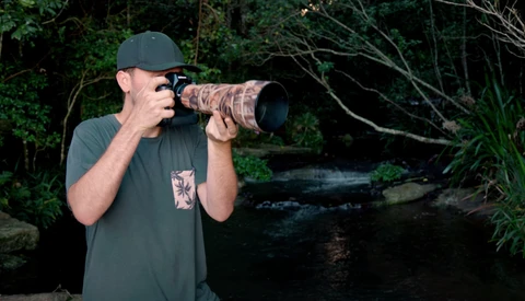 Photographer with telephoto lens in forest setting near water.