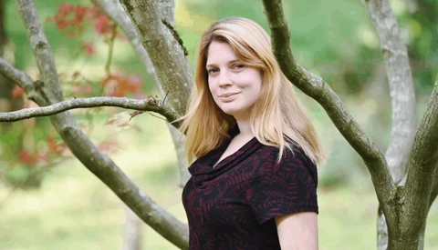 Young woman with blonde hair leaning against a tree branch in natural outdoor setting.
