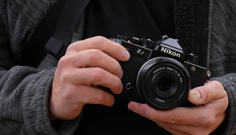Hands holding a black Nikon film SLR camera with a standard lens against a dark textured jacket.