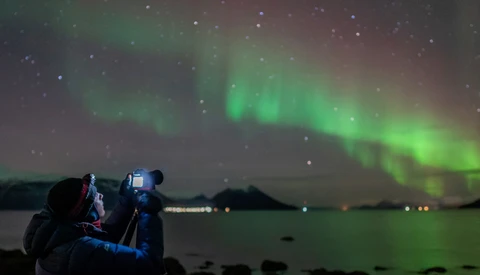 Photographer with camera on tripod capturing aurora borealis over dark water at night.