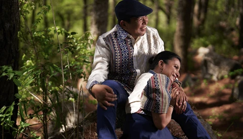 Adult and child in embroidered traditional clothing crouching together in a forested setting.
