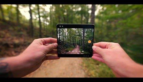 Hands holding a smartphone in landscape orientation displaying a forest trail in the camera viewfinder.