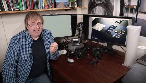 Photographer at desk with dual monitors displaying portfolio work and camera equipment.