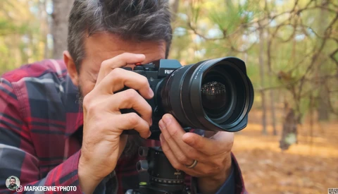 Photographer holding a camera with wide-angle lens, composing a shot in an autumn forest setting.