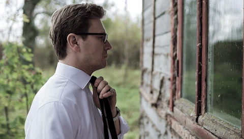 Man in white shirt and sunglasses looking toward weathered greenhouse window with green foliage backdrop.