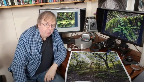 Photographer reviewing a forest landscape print at his desk workstation with dual monitors.