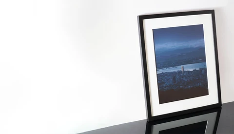 Framed photograph of a nighttime cityscape with illuminated buildings and water reflection displayed on a white wall.