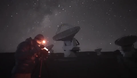 Radio telescope dishes silhouetted against a starry night sky with an explosion or bright light source illuminating one telescope in orange and red.