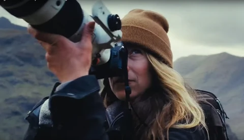 Photographer with long blonde hair wearing a tan beanie looks through a camera viewfinder while holding binoculars in a mountainous landscape.