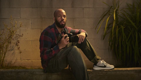 Photographer sitting against concrete wall holding camera, wearing plaid shirt and sneakers.
