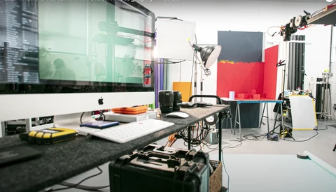 Professional photography studio workspace with Mac monitor, lighting equipment, and red backdrop panels.
