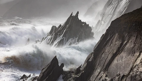 Dramatic coastal rock formations surrounded by crashing waves and sea spray in monochromatic tones.