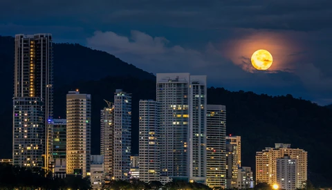 Illuminated high-rise buildings silhouetted against a full moon rising over mountains at dusk.