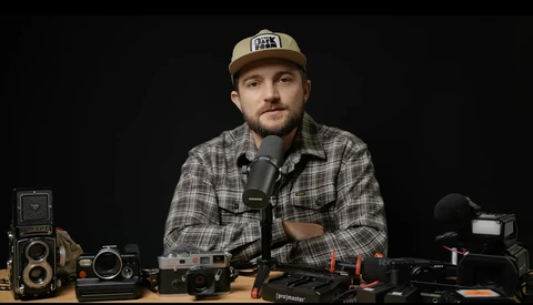Photographer at desk with vintage and modern cameras speaking into microphone against dark background.