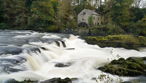 Flowing waterfall cascading over rocks with a stone mill building nestled among trees on the riverbank.