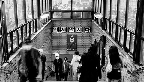 Blurred commuters moving through a subway station stairwell with Korean signage and brick walls.