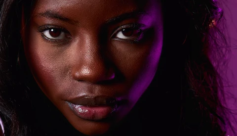 Close-up portrait of a woman with deep brown skin tone against a dark background with purple lighting.