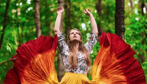 Woman with arms raised wearing flowing red and yellow fabric against green forest backdrop.