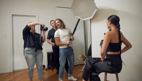 Photography studio setup with model posing on stool while crew adjusts octabox softbox lighting.
