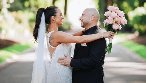 Bride and groom embrace outdoors while holding a bouquet of pink roses on their wedding day.