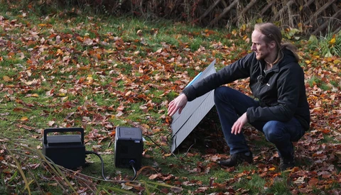 Photographer crouching beside solar panels on autumn ground covered in fallen leaves.