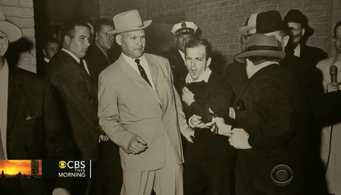 Men in suits and fedoras gathered in a brick corridor, one in a light tan coat being escorted by officials.