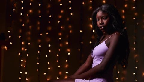 Woman in lavender tank top posed against warm bokeh lights in dark background.