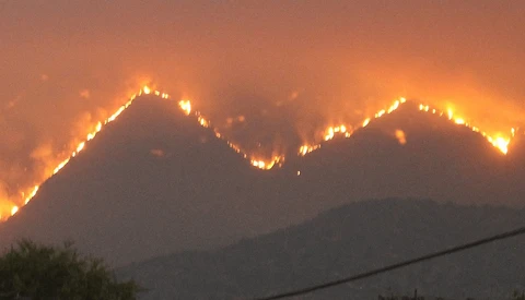 Wildfire spreading across a mountain ridge at night, with flames creating bright lines of fire against dark slopes and smoky sky.