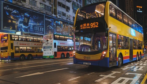 Double-decker buses on a Hong Kong street at night with illuminated buildings and advertisements in the background.