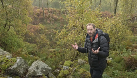 Man in black puffer jacket gesturing while speaking in a forested autumn setting.
