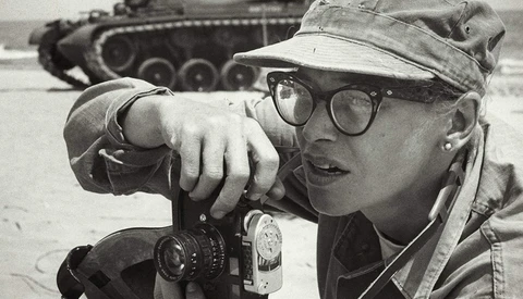 Photographer wearing glasses and cap looking through rangefinder camera with military tank blurred in background.