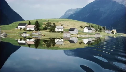 Alpine valley with scattered houses reflected in still lake water, surrounded by steep forested mountains.