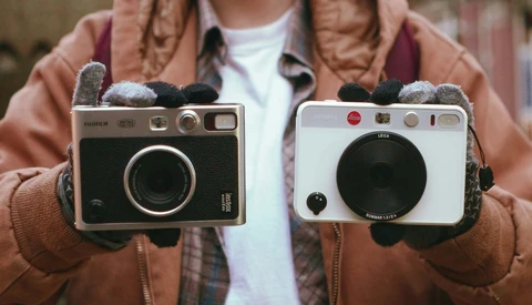 Person in brown jacket holding two vintage instant cameras side by side, wearing gray mittens.