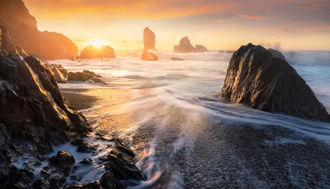 Rocky sea stacks silhouetted against golden sunrise over sandy beach with breaking waves.