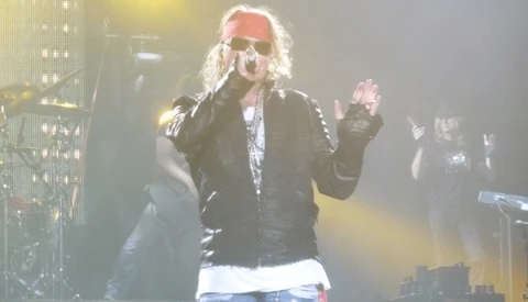 Performer on stage wearing sunglasses and leather jacket, singing into microphone with dramatic yellow stage lighting.