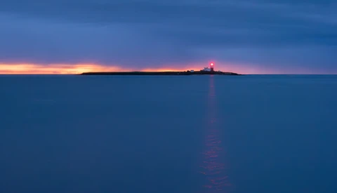 Lighthouse silhouetted on distant horizon at twilight with calm water and golden sunset reflection.