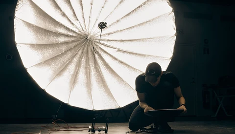 Photographer crouching beside a large parabolic reflector on a dark studio floor.
