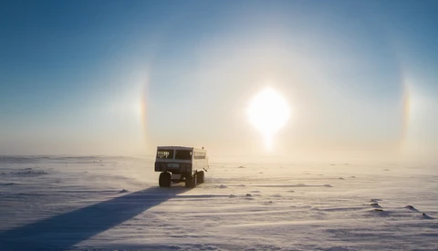 Military truck driving across a frozen landscape with sun dogs visible in the bright sky.