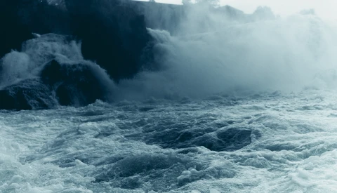 Powerful waves crash against dark rocky cliffs shrouded in white sea spray and mist.