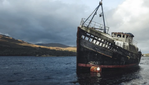 Abandoned ship with deteriorating hull tilted in shallow water against cloudy sky and barren hillside.